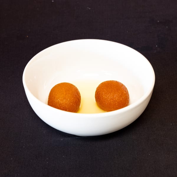 Two golden-brown gulab jamun, Indian fried dough balls, soaking in syrup in a white bowl.